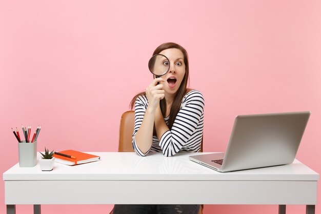 Mulher curiosa em roupas casuais, olhando através da lupa, senta-se no trabalho na mesa branca com o laptop pc contemporâneo isolado no fundo rosa pastel. Conceito de carreira empresarial de realização. Copie o espaço.