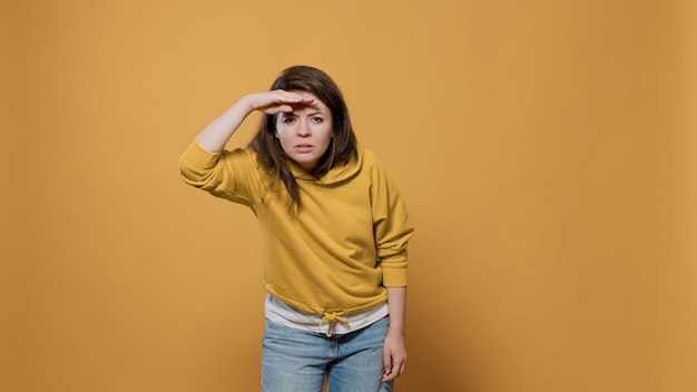 Mulher curiosa olhando para longe procurando algo com a palma da mão na testa no estúdio. Jovem explorando o horizonte à procura de alguém sobre fundo amarelo.