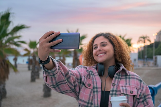 Mulher curvilínea com cabelos cacheados tira uma selfie com seu telefone na costa ao pôr do sol