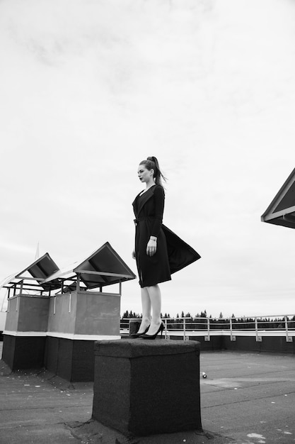 Foto mulher da cidade grande com rabo de cavalo de pé no topo do telhado em um vestido preto soprado pelo vento. preto e branco.