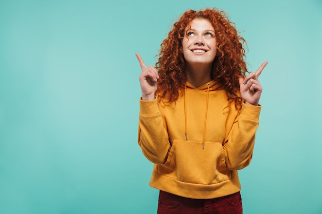 Mulher de 20 anos satisfeita com cabelo ruivo cacheado sorrindo e apontando o dedo para copyspace isolado na parede azul