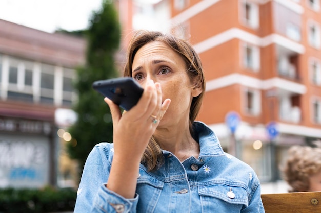 Mulher de 40 anos na rua enviando mensagem de voz com o smartphone ou consultando o assistente de voz