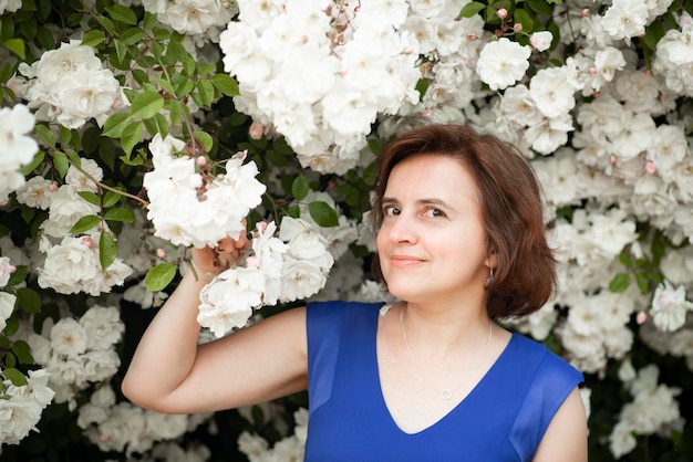 Mulher de 48 anos em um vestido azul perto de uma rosa branca florescendo