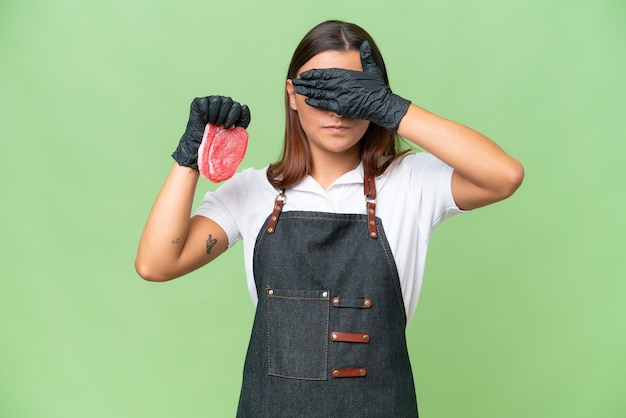 Mulher de açougueiro vestindo um avental e servindo carne fresca isolada em fundo croma verde cobrindo os olhos pelas mãos Não quer ver algo