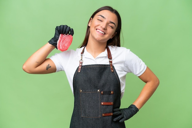 Mulher de açougueiro vestindo um avental e servindo carne fresca isolada em fundo croma verde posando com os braços no quadril e sorrindo