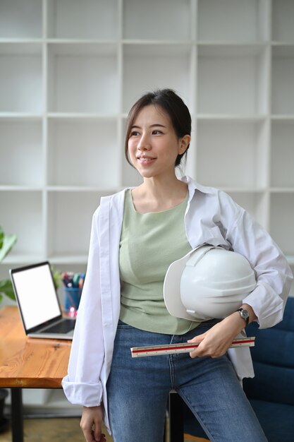 Mulher de arquiteto confiante segurando com capacete de segurança e permanente no escritório.