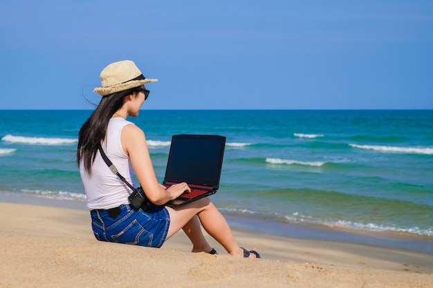 Mulher de Ásia, sentado na praia e usando o laptop para o seu trabalho e verificação de negócios durante as férias.