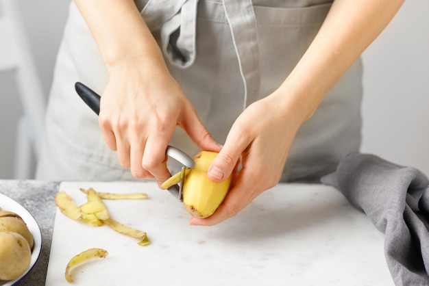 Mulher de avental descascando batata na placa de mármore Fundo branco Mãos descascando batata com descascador de cozinha Preparação de alimentos