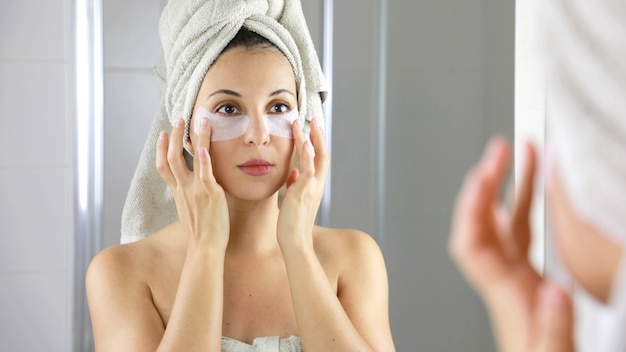 Mulher de beleza aplicando máscara antifadiga sob os olhos, olhando-se no espelho no banheiro.