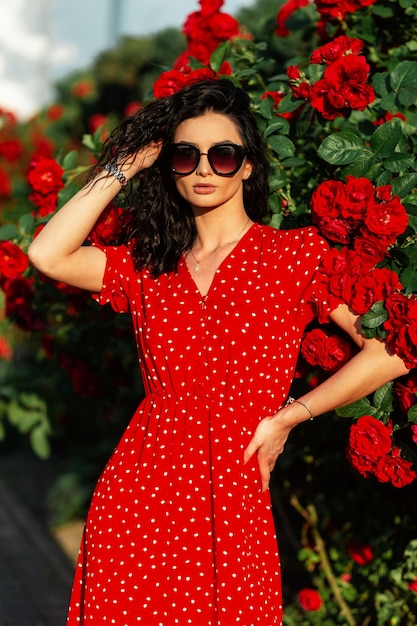 Mulher de beleza elegante com óculos de sol da moda em um vestido vermelho da moda fica perto de rosas vermelhas no parque