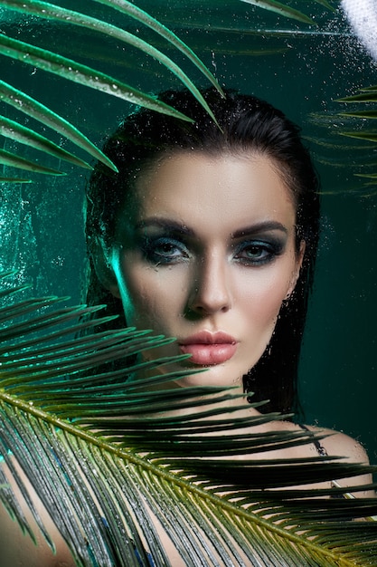 Mulher de beleza em folhas de palmeira molhada maquiagem, menina retrato tropical em maiô verde em ramos de palmeira em estúdio, fumaça e gotas de chuva no vidro. Mulher sexy com maquiagem verde brilhante