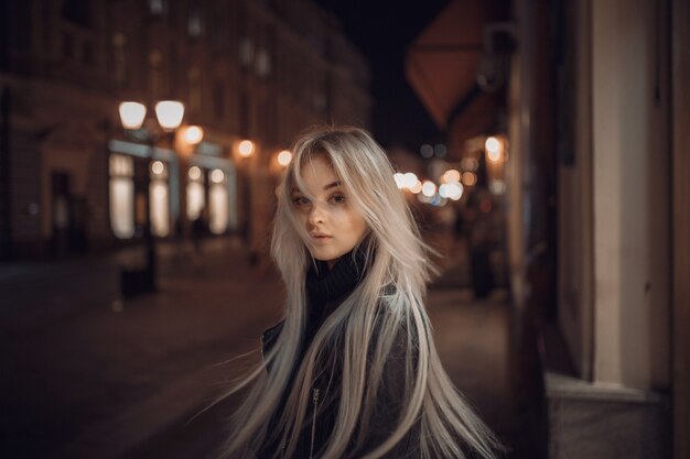 Foto mulher de beleza jovem sexy posando sobre fundo de rua iluminada cidade à noite. férias de férias. jovem loira elegante com cabelo comprido