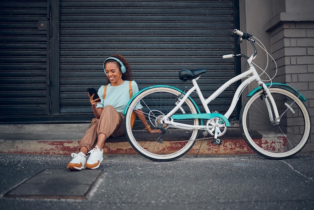 Foto mulher de bicicleta e fone de ouvido com telefone na cidade relaxe com bicicleta e mensagens de texto felizes aplicativos de mídia social e bate-papo menina senhora e com transporte ecológico moderno ou visual moderno, sente-se na calçada