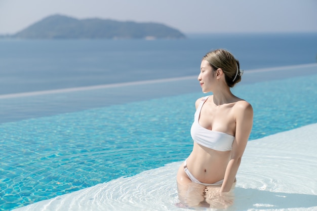 Mulher de biquíni branco relaxando na beira de uma piscina infinita com vista para o mar azul.