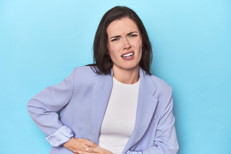 Foto mulher de blazer azul sobre fundo azul com dor de estômago e dor no fígado