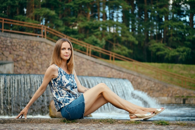 Mulher de blusa sem mangas e saia jeans, descansando na área de recreação do país, sentada perto de cachoeira artificial