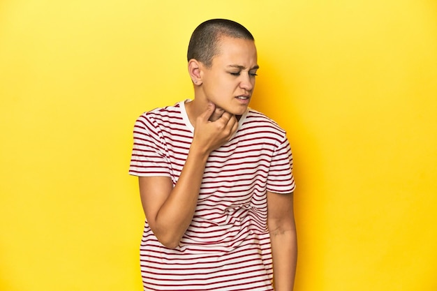 Mulher de cabeça rapada com camiseta listrada vermelha com fundo amarelo sofre dor na garganta devido a um vírus ou infecção