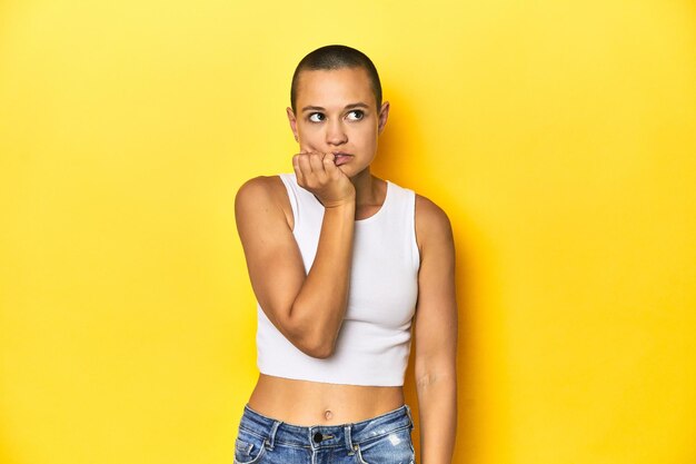 Mulher de cabeça raspada em top branco pano de fundo amarelo roendo unhas nervosas e muito ansiosas