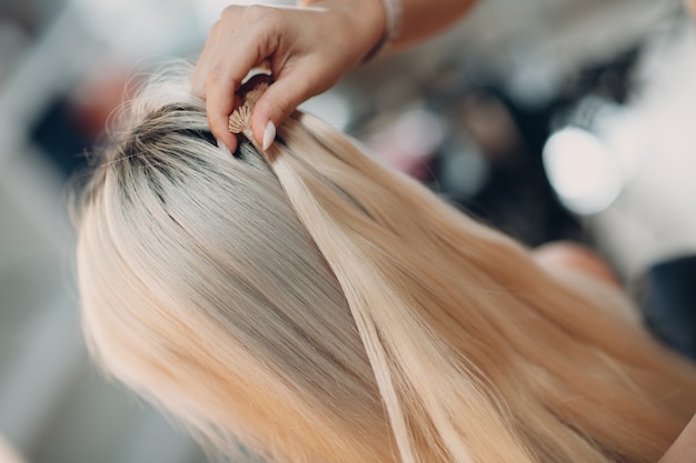 Mulher de cabeleireiro fazendo extensões de cabelo para jovem loira em salão de beleza. Extensão de cabelo profissional.