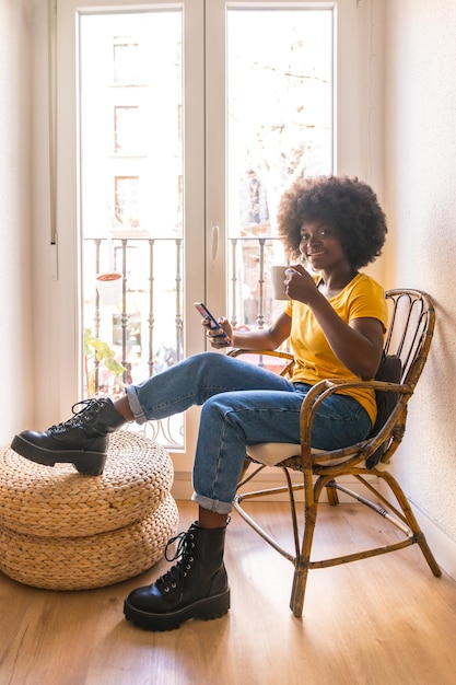 Mulher de cabelo afro na janela de casa tomando uma bebida quente café da manhã olhando para o celular