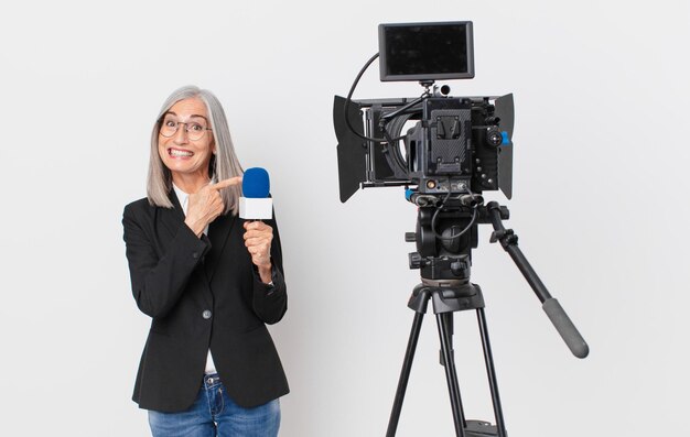 Mulher de cabelo branco de meia-idade olhando animada e surpresa apontando para o lado e segurando um conceito de apresentador de televisão de microfone