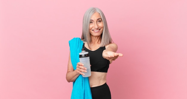 mulher de cabelo branco de meia-idade, sorrindo alegremente com simpáticos e oferecendo e mostrando um conceito com uma toalha e uma garrafa de água. conceito de fitness