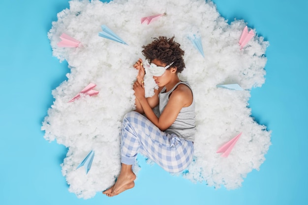 Mulher de cabelo encaracolado calma serena vê bons sonhos depois de um dia de trabalho usa pijama e máscara de dormir nos olhos posa na nuvem branca com aviões de papel ao redor isolado sobre fundo azul hora de dormir