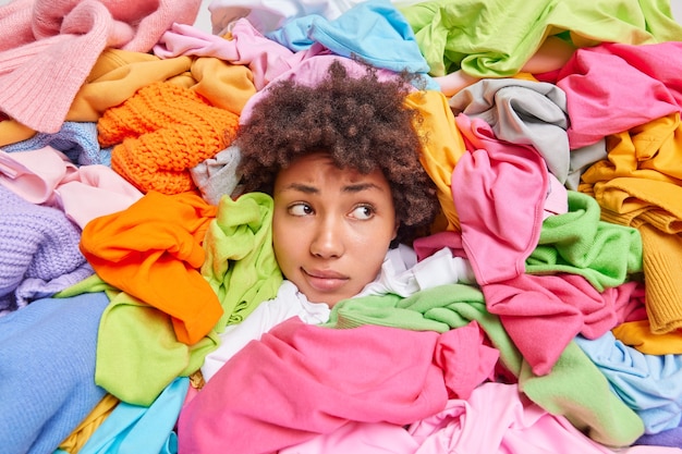 Mulher de cabelo encaracolado coleta itens de roupas em boas condições para loja de consignação ou brechó cercada por enormes pilhas de roupas multicoloridas focadas em uma expressão de cansaço. Reciclagem de têxteis