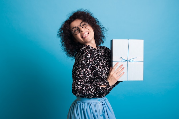 Mulher de cabelo encaracolado com óculos e vestido apresentando para a câmera uma caixa com um presente posando na parede azul do estúdio