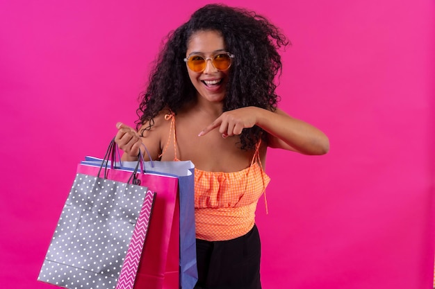 Mulher de cabelo encaracolado em um conceito de compras de fundo rosa apontando para o tiro de estúdio de sacos de papel