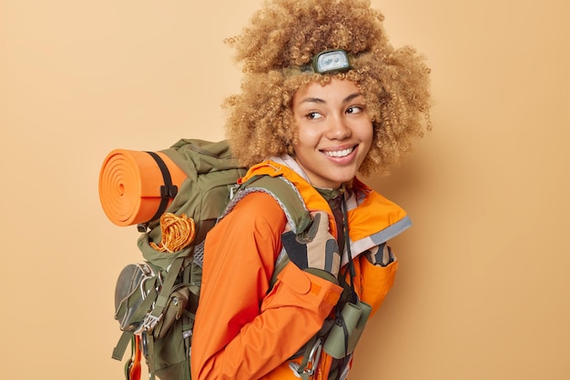 Foto mulher de cabelo encaracolado feliz tem caminhada tem bom humor durante o tempo de recreação carrega mochila usa farol de blusão para olhar na escuridão isolada em fundo marrom alpinista feminina caminhadas na floresta