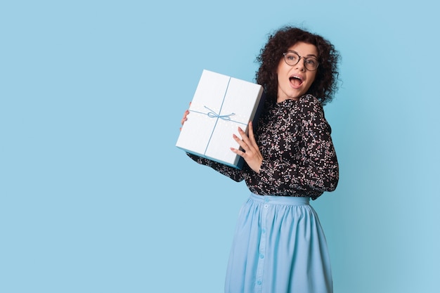 Mulher de cabelo encaracolado mostrando na frente uma caixa de presente posando feliz perto do espaço azul