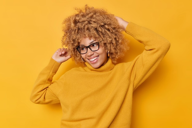 Mulher de cabelo encaracolado positiva com expressão alegre se move ativamente com os braços levantados, danças internas e clima otimista, vestida com um casaco casual quente isolado sobre fundo amarelo. Foto monocromática