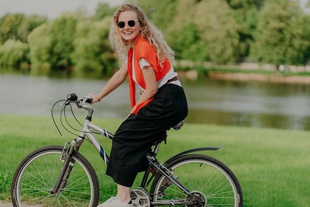 Mulher de cabelo encaracolado satisfeita de aparência agradável passa o tempo livre em passeios ao ar livre, bicicleta passa lazer sozinho desfruta do frescor da natureza Olha para a câmera com um sorriso agradável Horário de verão