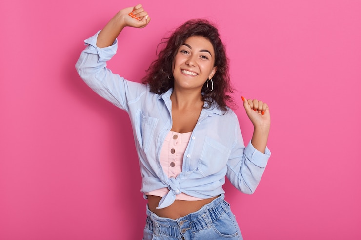 Foto mulher de cabelo escura feliz dançando contra parede rosada, levantando as mãos, vestindo jeans e camisa azul, olhando sorrindo expressando emoções positivas.