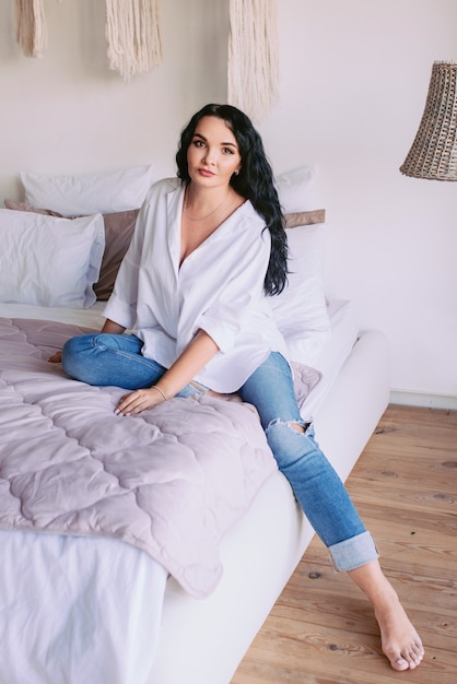 Foto mulher de cabelo escuro em camisa branca e jeans sentada triste em sua cama