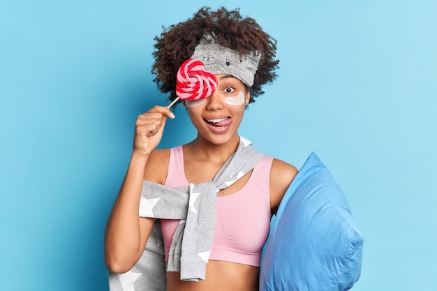 Mulher de cabelo muito encaracolado cobre os olhos com um doce lambe os lábios vestida com roupas casuais aplica adesivos de colágeno antes de dormir segura o travesseiro isolado na parede azul