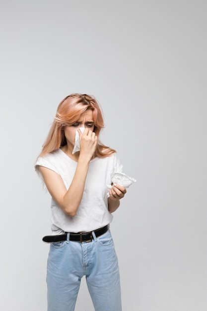 Foto mulher de cabelo rosa e camisa branca sopra o nariz num lenço possivelmente com sintomas de resfriado