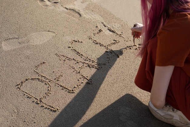 mulher de cabelo rosa na praia com um pedaço de pau na areia escreve a palavra paz