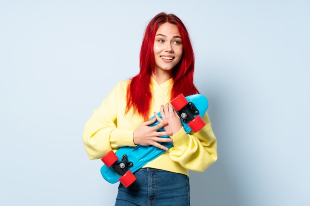 Mulher de cabelo vermelho adolescente isolada na parede azul com um skate com expressão feliz