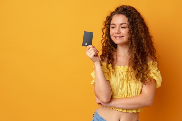 Foto mulher de cabelos cacheados segurando um cartão de crédito preto sobre fundo amarelo