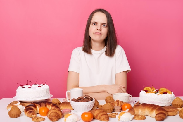 Mulher de cabelos castanhos chateada e descontente confusa em camiseta branca sentada à mesa com massa olhando para a câmera com rosto triste mantém dieta isolada sobre fundo rosa