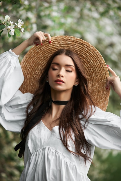 Mulher de cabelos castanhos com um grande chapéu de palha e um vestido branco posa no contexto de árvores brancas florescendo. Aparência romântica, beleza natural, pele facial limpa