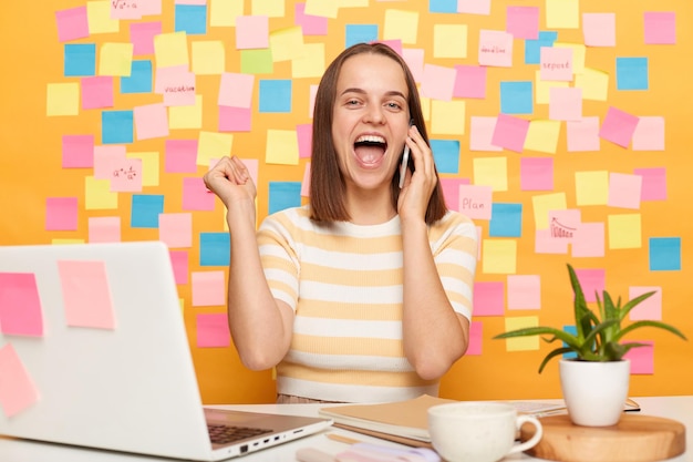 Mulher de cabelos castanhos extremamente feliz vestindo camiseta listrada sentada à mesa em seu local de trabalho com laptop contra parede amarela com notas adesivas falando telefone punhos cerrados gritando alegremente