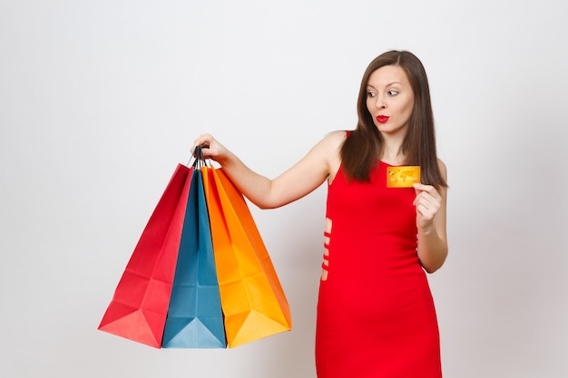 Mulher de cabelos castanhos jovem elegante glamour atraente vestida de vermelho segurando o cartão de crédito, pacotes coloridos com compras após as compras, isoladas no fundo branco. Copie o espaço para anúncio.