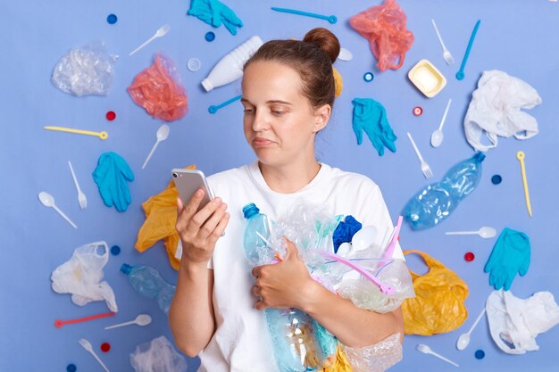 Mulher de cabelos castanhos vestindo camisa branca isolada na parede azul com lixo ao redor usando telefone celular lendo mensagens nas redes sociais tendo uma pausa enquanto limpava a rua do lixo