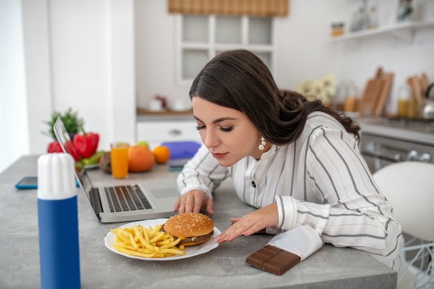 Mulher de cabelos escuros em uma blusa listrada comendo hambúrguer com batata frita