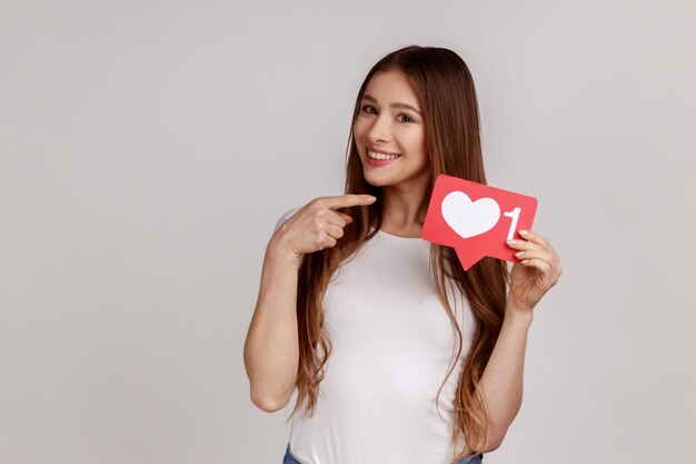 Foto mulher de cabelos escuros sorridente, apontando o dedo para o sinal com contador, blogueiro esperando feedback e taxa positiva, vestindo camiseta branca. tiro de estúdio interior isolado em fundo cinza.