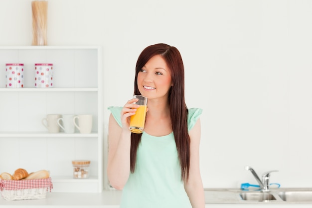 Mulher de cabelos ruivos bem-vinda que aprecia um copo de suco de laranja na cozinha