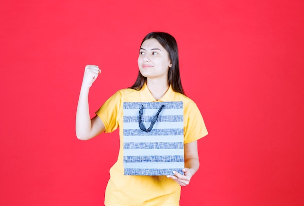 Mulher de camisa amarela, segurando uma sacola de compras azul e mostrando sinal positivo com a mão.